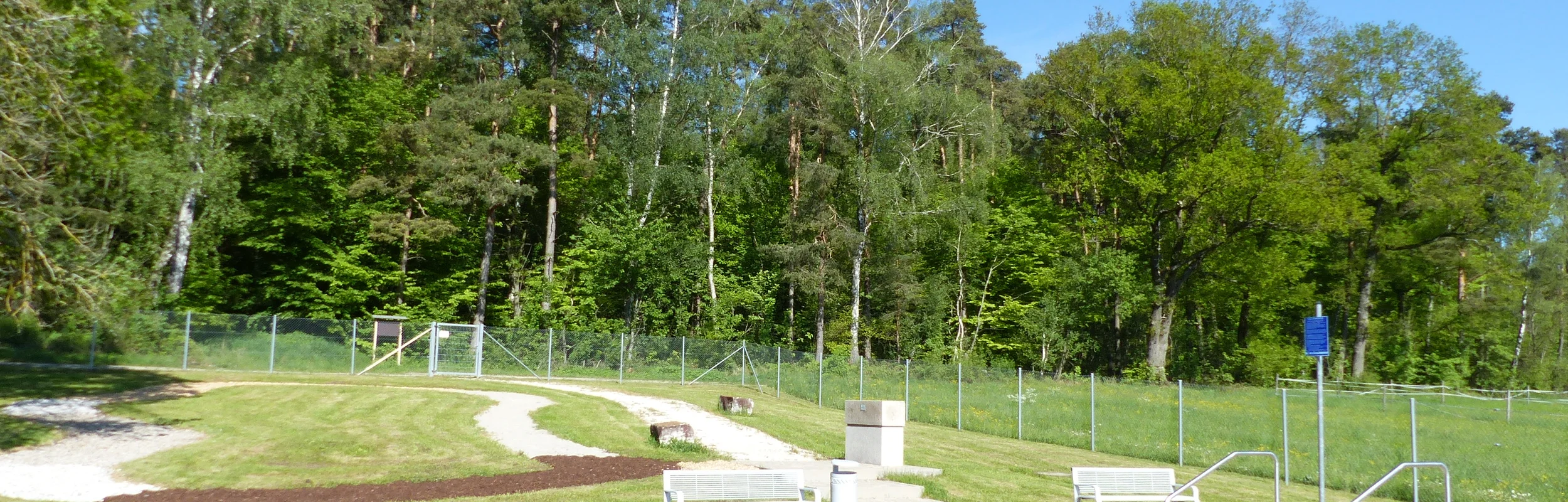 Rundes Kneippbecken in Monheim mit einem Geländer und einem Barfußpfad auf einer Wiese, umgeben von einer großen Grünfläche und Bäumen und Sitzmöglichkeiten. Ein Zaun im Hintergrund.