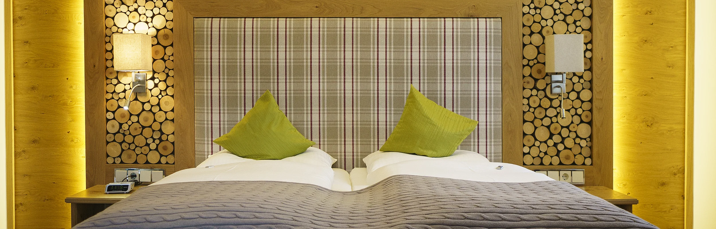 Wir sehen  das gemütliche Doppeltbett aus Holz. Das Bett ist gemacht und grüne Dekokissen geben etwas Farbe. Dahinter ist das Kopfende mit kariertem Stoff zu sehen. Die Wand hinter dem Bett ist zu dreiviertel mit abgeschnittenen runden Holzscheiben, die mit einem Rahmen, der beleuchtet ist,  eingefasst wurden. Die restliche Wand ist mit Holzbrettern verkleidet. Links ist eine Öffnung im Raum zu sehen. Zudem sieht man die weiße Decke mit der Lampe, die ebenfalls leuchtet.