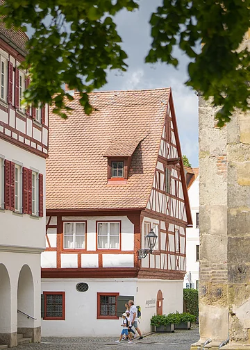 Fachwerkhaus mit rotem Dach und weißen Wänden, umgeben von Bäumen. Eine Familie geht auf einem gepflasterten Weg vorbei.