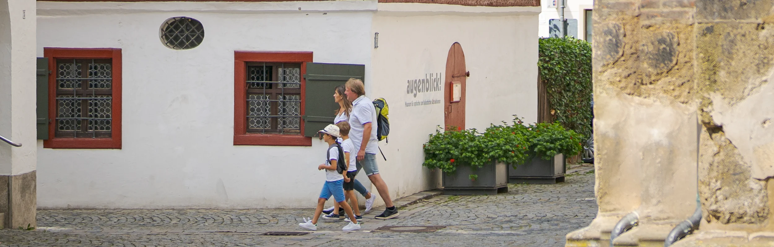 Fachwerkhaus mit rotem Dach und weißen Wänden, umgeben von Bäumen. Eine Familie geht auf einem gepflasterten Weg vorbei.
