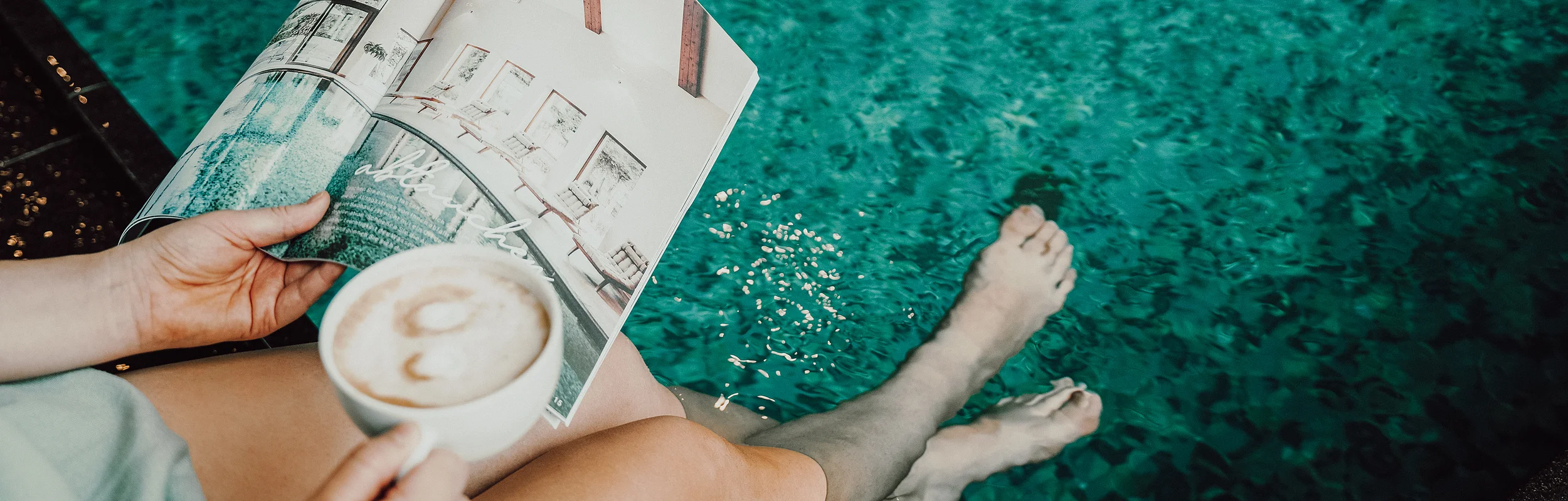 Person hält eine Tasse Kaffee und ein Magazin, Füße im Wasser.