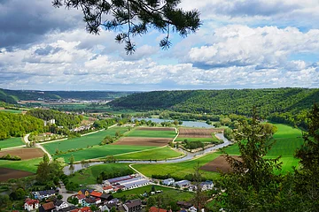 Landschaft mit Feldern, Fluss und Waldhügeln unter bewölktem Himmel, im Vordergrund ein Dorf.