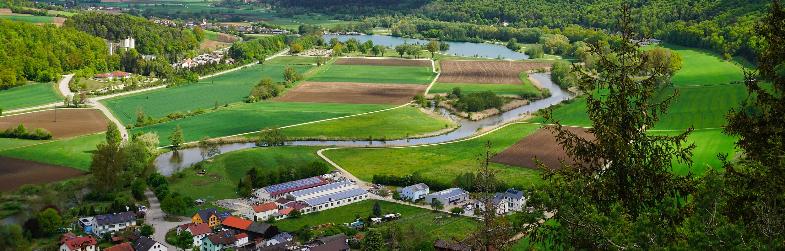 Landschaft mit Feldern, Fluss und Waldhügeln unter bewölktem Himmel, im Vordergrund ein Dorf.