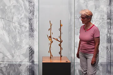 Original und Rekonstruktion des Kultbäumchens aus Manching Eine Person betrachtet eine Skulptur mit goldfarbenen Zweigen und Blättern in einer gläsernen Vitrine.