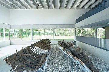 Die Schiffshalle mit den römischen Booten aus Oberstimm Zwei antike Holzbootwracks auf Kiesboden in einem modernen Ausstellungsraum mit Glaswänden.