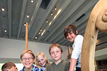 Kinder erkunden die Schiffshalle des kelten römer museums Fünf Kinder stehen in einem hölzernen Boot in einem Gebäude mit Betonwänden und Deckenbeleuchtung.