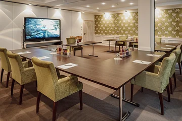 Tagungen Hotel Eisvogel Tische Konferenzraum mit U-förmigem Tisch, grünen Stühlen, einem großen Bildschirm und Getränken auf dem Tisch.