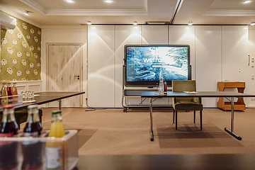 Tagungen Hotel Eisvogel innenraum Konferenzraum mit einem großen Bildschirm, einem Tisch mit Getränken und Stühlen, florale Tapete an der Wand.