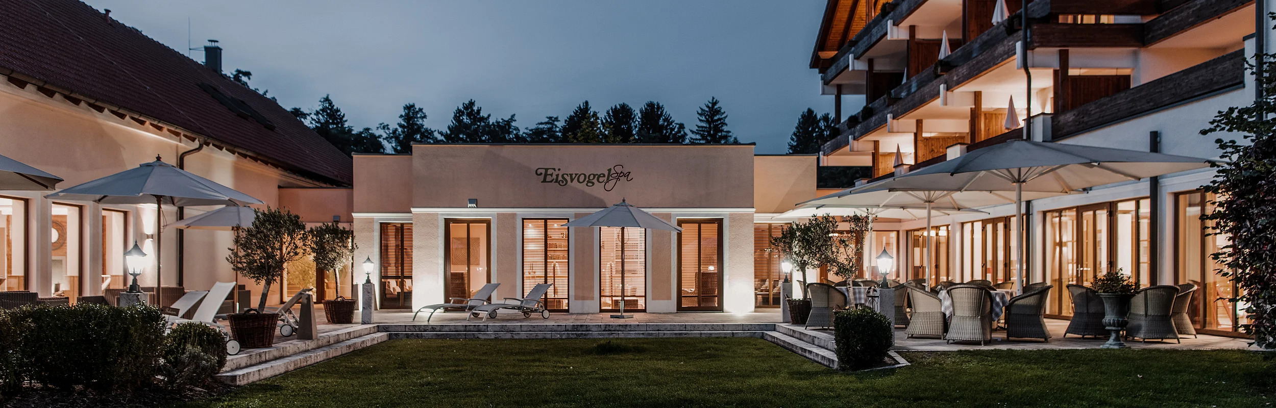 Hotel Eisvogel Außenansicht Außenansicht eines beleuchteten Gebäudekomplexes mit Terrasse und Garten bei Dämmerung.