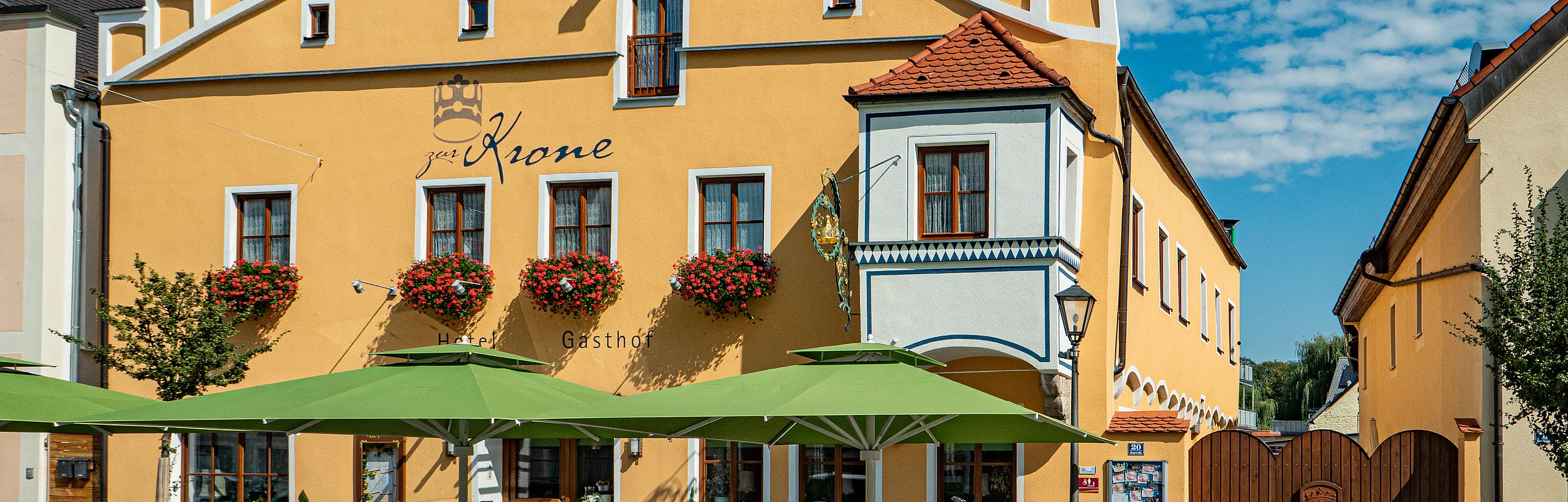 Gelbes Gebäude mit Ziergiebel, Fensterläden und Blumenkästen. Davor Tische mit grünen Sonnenschirmen auf einem gepflasterten Platz.