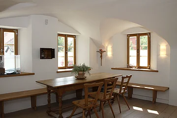 Esszimmer mit Holztisch, Stühlen und Bänken. Zwei Fenster, weiße Wände, Pflanze auf dem Tisch.