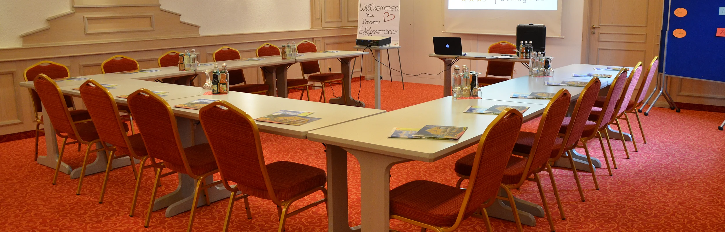 Konferenzraum mit U-förmiger Tischanordnung, roten Stühlen, Flipchart und Projektor.