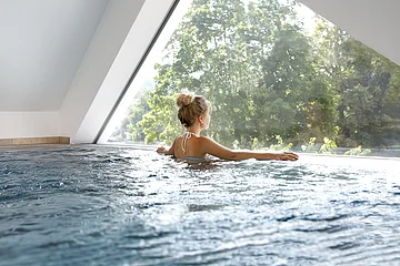 Ein Gast genießt den Ausblick von Schwimmbecken durch ein Fenster nach draußen in die Naturlandschaft.
