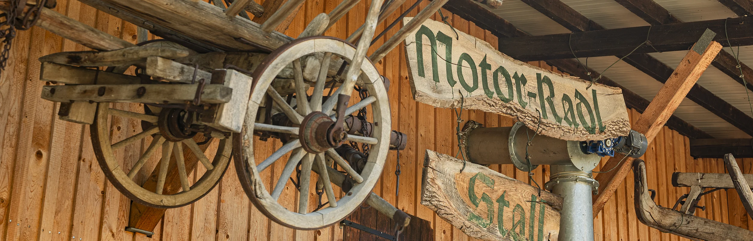 Holzwagenrad und Holzschilder mit der Aufschrift "Motor-Radl Stall" an einer Holzwand.