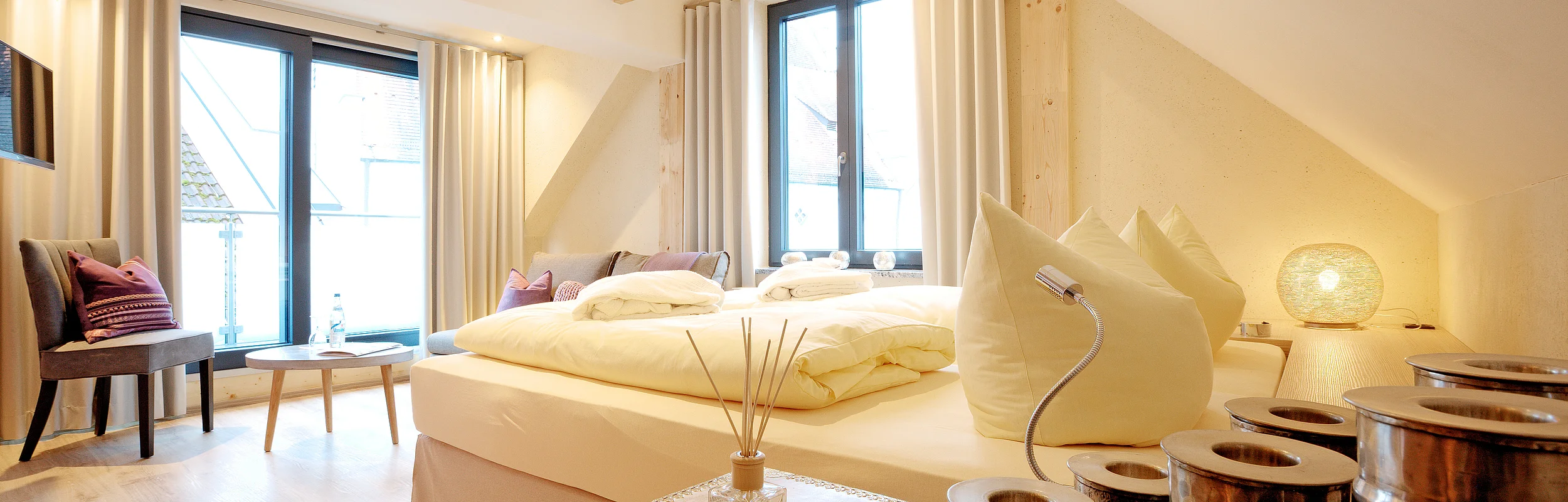 Idylle zu zweit Hotel Die Gams Helles Schlafzimmer mit Doppelbett, großen Fenstern, Holzboden und dekorativen Kissen. Ein Tisch mit Stühlen steht am Fenster.