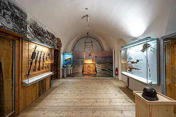 Ein Museumsraum mit Vitrinen, die historische Waffen und Ausrüstungsgegenstände zeigen. Ein Helm steht auf einem Holzpodest.