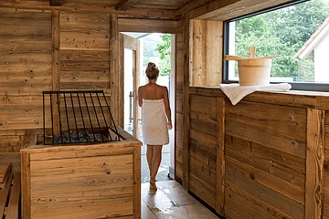 Hotel die Gams Beilngries Eine Person in einem Handtuch verlässt eine Holzsauna durch eine Tür. Ein Saunaeimer steht auf einem Handtuch am Fenster.