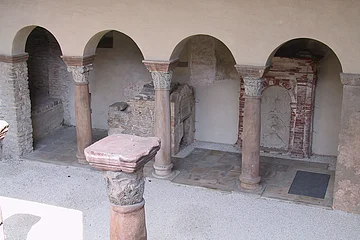 Sola-Basilika Steinarkaden mit verzierten Säulen in einem historischen Gebäude. Im Hintergrund sind Mauerreste sichtbar.