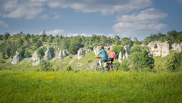 Zwei Personen fahren mit Fahrrädern auf einem grasbewachsenen Feld vor einer Felslandschaft.