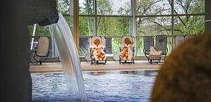 Innenpool mit Wasserfall, zwei Personen in weißen Bademänteln auf Liegestühlen, große Fenster mit Blick auf Bäume.