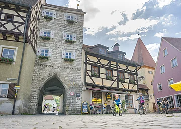 Kinder fahren mit Fahrrädern auf einem gepflasterten Platz vor Fachwerkhäusern und einem Torbogen.