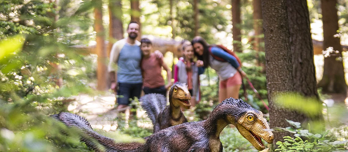 Familie im Dinosauriermuseum in Denkendorf Zwei Dinosauriermodelle im Wald, unscharf im Hintergrund eine Gruppe von Menschen.
