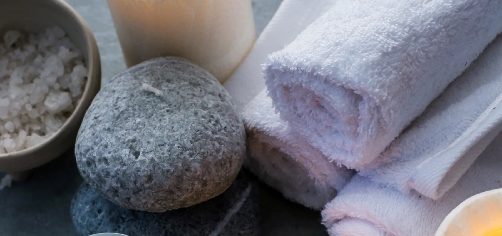 Kuscheltage im Landhotel Zur Jurahöhe Kerzen, Handtücher, Steine, ein Glas mit Creme, eine Schale mit Salz und ein Holzlöffel auf grauem Hintergrund.
