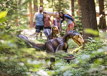 Familie im Dinosauriermuseum in Denkendorf Zwei Dinosauriermodelle im Wald, unscharf im Hintergrund eine Gruppe von Menschen.