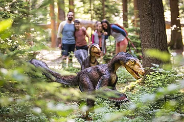 Familie im Dinosauriermuseum in Denkendorf Zwei Dinosauriermodelle im Wald, unscharf im Hintergrund eine Gruppe von Menschen.
