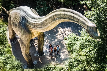 Familie im Dinosauriermuseum in Denkendorf Große Dinosaurierstatue im Wald, Menschen stehen darunter und schauen nach oben.