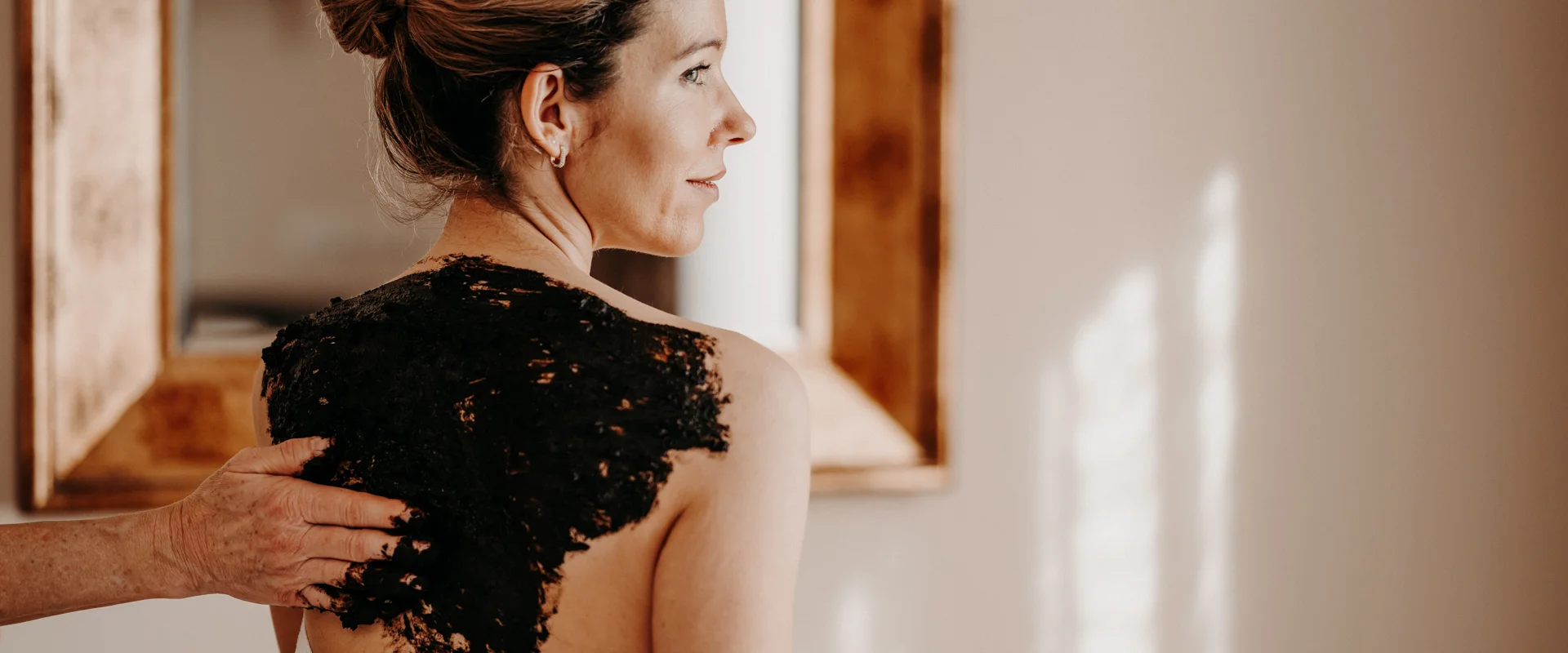 Person mit hochgestecktem Haar bekommt eine schwarze Paste auf den Rücken aufgetragen.