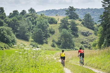 Zwei Personen fahren mit Fahrrädern auf einem Pfad durch eine grüne, hügelige Landschaft.