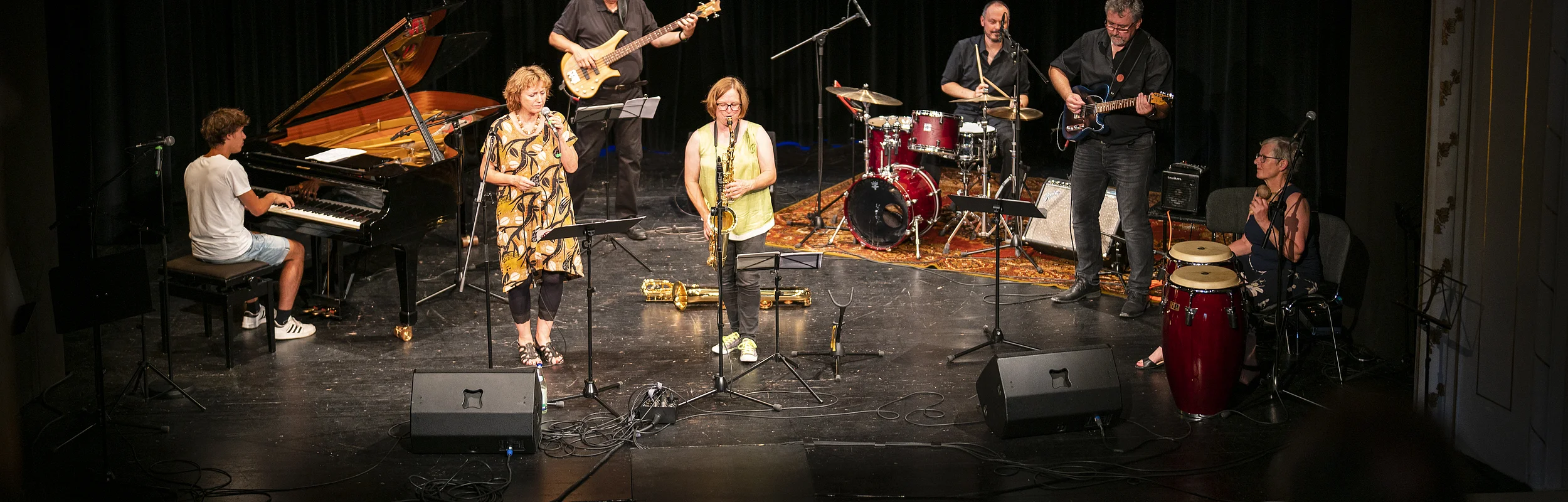 Eine sechsköpfige Band spielt auf einer Bühne. Ein Pianist, Sängerin, Saxophonistin, Bassist, Schlagzeuger und Gitarrist sind zu sehen.
