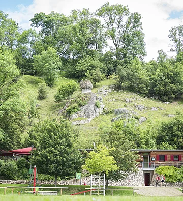 Grüne Hügellandschaft mit Felsen, Bäumen und einem Gebäude im Vordergrund. Spielplatz mit Klettergerüst sichtbar.