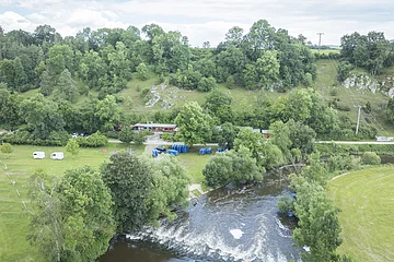 Landschaft mit Fluss, Bäumen, Hügeln und einem Gebäude. Wohnwagen und blaue Boote auf einer Wiese.