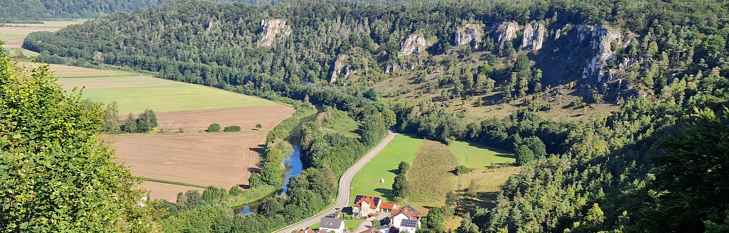 Landschaft mit Fluss, Feldern, Wald und kleinem Dorf im Tal. Hügel im Hintergrund.