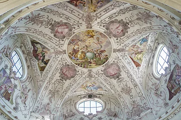 Deckenfresko mit religiösen Szenen und Ornamenten in einer Kirche, umgeben von Fenstern und Stuckverzierungen.