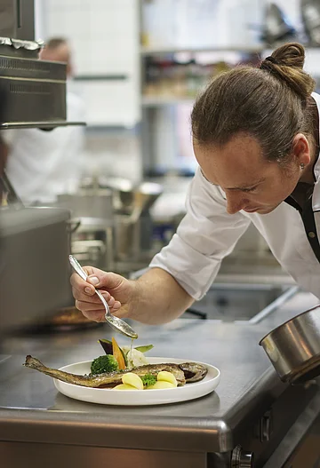 Ein Koch in einer Küche verfeinert ein Fischgericht auf einem Teller mit einer Soße.