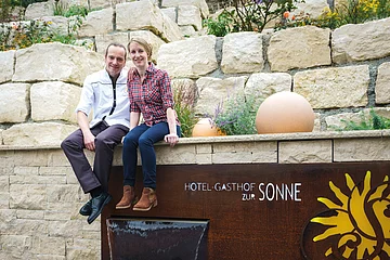 Zwei Personen sitzen auf einer Steinmauer neben einem Schild mit der Aufschrift "Hotel-Gasthof zur Sonne".