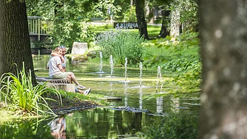 Personen sitzen auf einer Bank am Teich mit Springbrunnen, umgeben von Bäumen und Grünpflanzen.