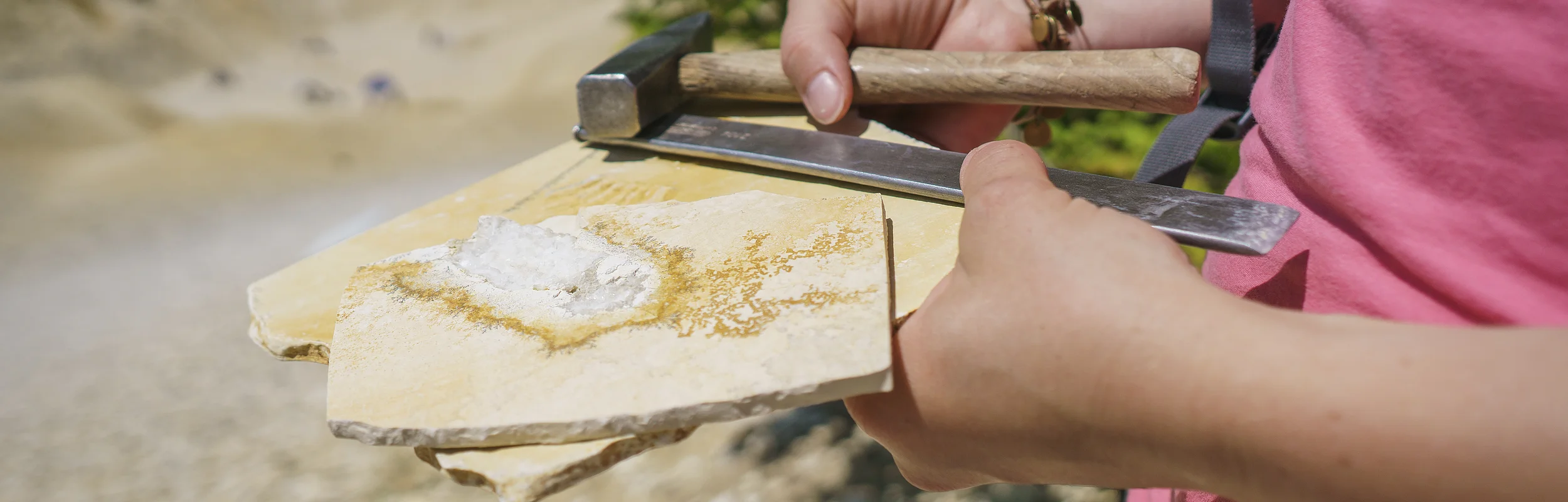 Person hält Hammer, Meißel und Gesteinsplatten mit einem weißen Mineral in einer Steinbruchumgebung