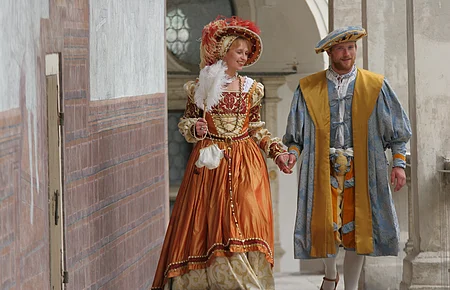 Paar in historischer Renaissancekleidung hält Händchen und geht durch einen Säulengang.