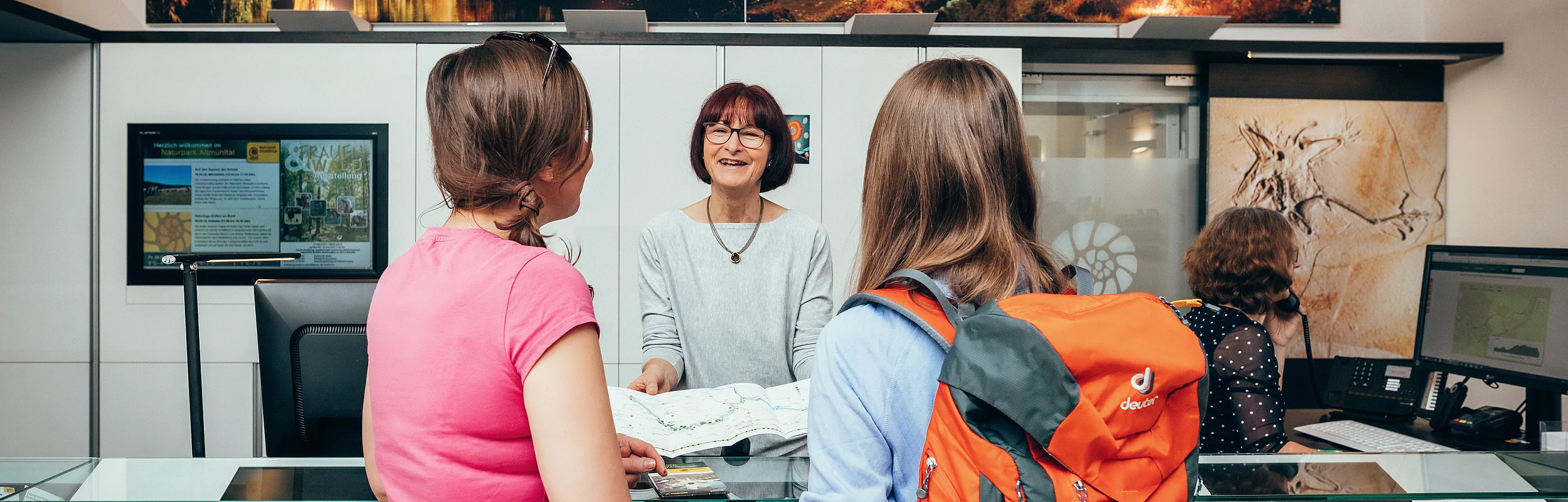Zwei Frauen mit Rucksack und pinkem Shirt erhalten an einem Informationsschalter eine Karte von einer Mitarbeiterin.