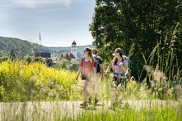 Drei Wanderer mit Rucksäcken auf Weg vor gelben Blumen und Dorf mit Kirchturm im Hintergrund