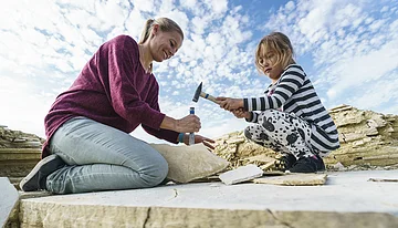 Frau und Mädchen schlagen mit Hammer und Meißel auf Steinplatte bei Ausgrabung im Freien ein.