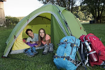 Zwei Personen liegen in einem grünen Zelt, davor Wanderschuhe und zwei große Rucksäcke auf Gras.