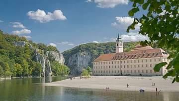 Flussufer mit Kiesstrand, altes Gebäude mit Turm und bewaldete Felsen unter blauem Himmel mit Wolken