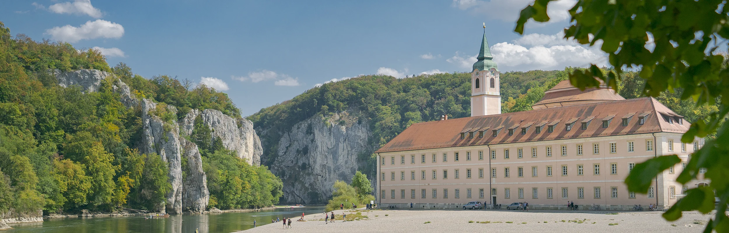 Flussufer mit Kiesstrand, altes Gebäude mit Turm und bewaldete Felsen unter blauem Himmel mit Wolken