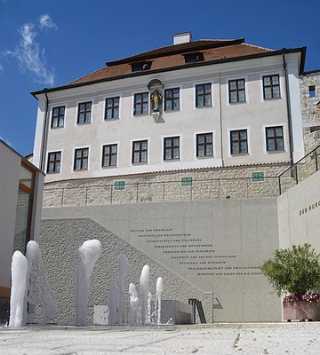Wasserspiele vor einer historischen Fassade mit Statue und Text an einer Betonwand bei klarem Himmel.