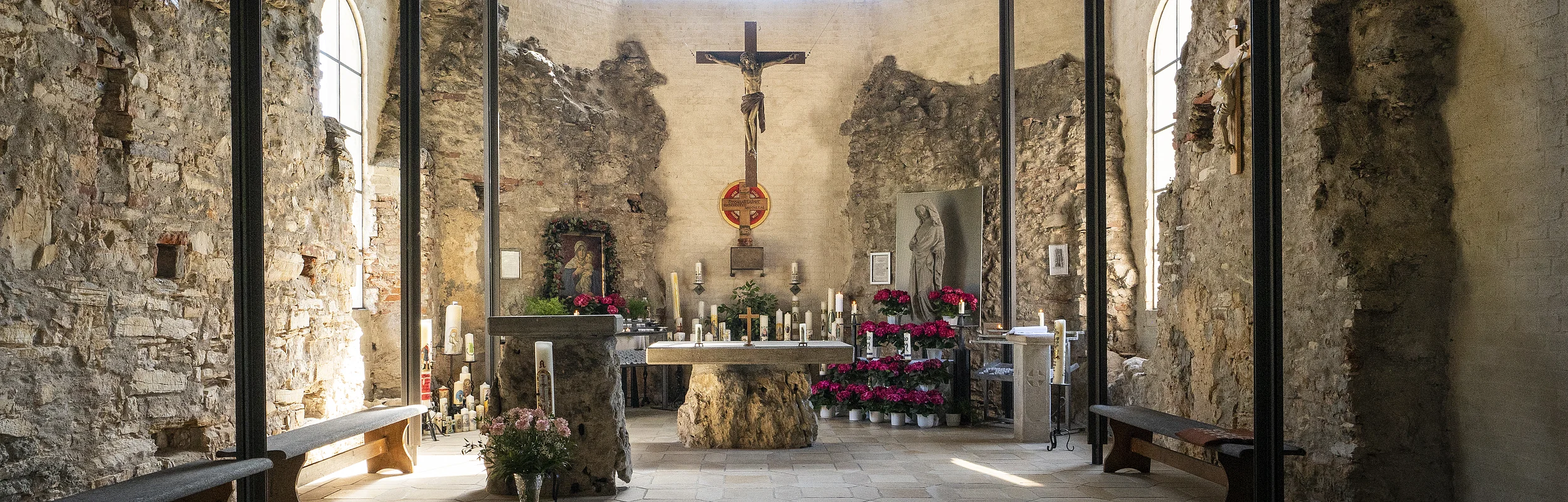 Innenraum einer Kapelle mit Altar, Kreuz, Kerzen und Blumen, umgeben von alten Steinmauern und Holzdecke